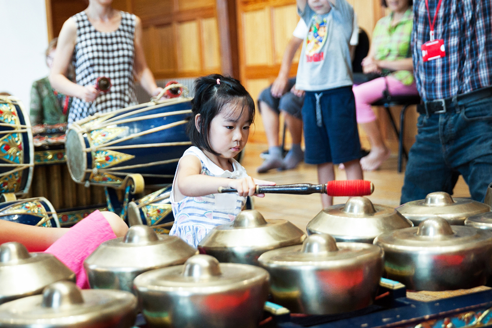 A young Asian girl playing the metal percussion instruments with a mallet, with a women playing a drum in the background.